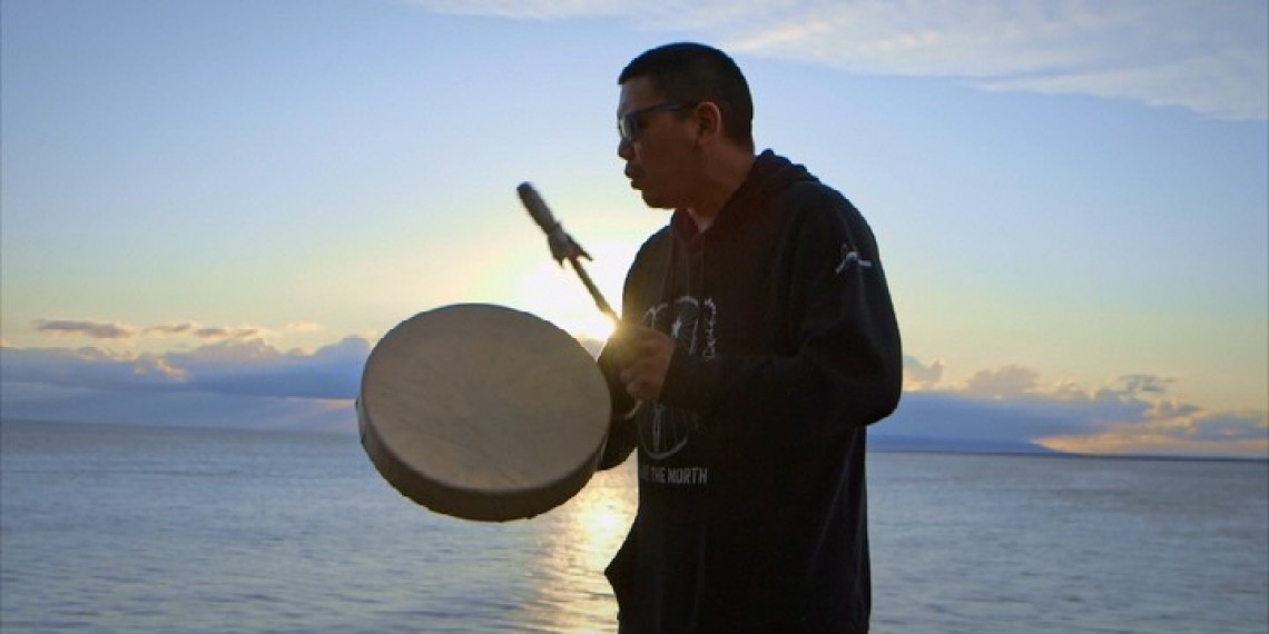 silhouette of man using Native Amercian drum in front of water at sunrise/sunset
